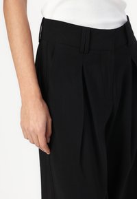 Black trousers featuring a smooth texture, pleated front design, and a fitted waistband, showcasing a flat pocket detail on the side.