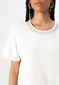 T-shirt en coton blanc avec un col ras du cou orné de perles argentées et de délicates accents en rhinestones autour du col. Manches courtes.