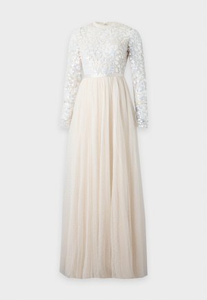 Femme portant une longue robe crème avec un corsage floral à sequins et des manches longues transparentes à pois, debout devant un fond blanc uni.