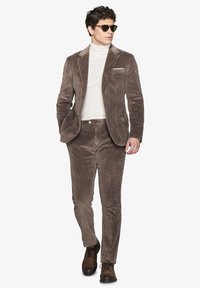 Brun fløjlssuit med en tekstureret overflade, matchende bukser og en lys højhalset sweater. Inkluderer solbriller og brune lædersko.