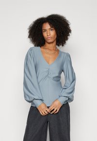 Blusa azzurra chiara con scollo a V, dettaglio frontale arricciato e maniche a palloncino voluminose. Tessuto liscio con una leggera brillantezza. Abbinata a jeans scuri.