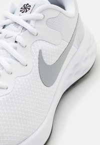Biały but sportowy z szarymi akcentami, chropowatą siateczkową cholewką, okrągłymi sznurowadłami, wyraźnym szarym logo Nike swoosh, amortyzowanym śródstopiem oraz czarną podeszwą zewnętrzną.