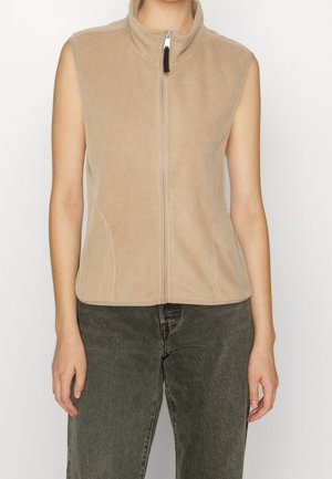 Veste en polaire beige sans manches avec fermeture éclair, portée par une personne avec un pantalon gris foncé, montrée du cou à la taille sur un fond uni.