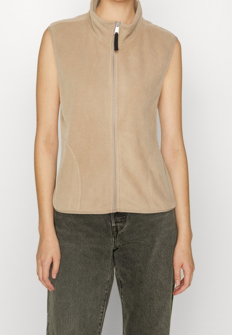 Veste en polaire beige sans manches avec fermeture éclair, portée par une personne avec un pantalon gris foncé, montrée du cou à la taille sur un fond uni.