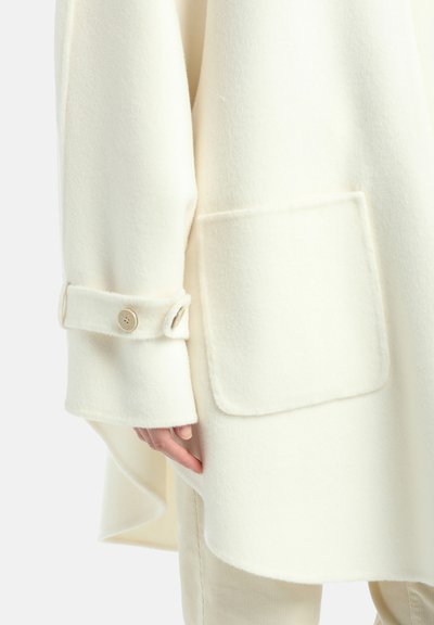 Witte wollen jas met een afgeronde zoom, grote zak en knoopdetail bij de mouwen. Zachte textuur, losse pasvorm en minimalistisch design.