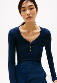 Bluză lungă dintr-un material côtelat, de culoare bleumarin, cu decolteu adânc în V și trei nasturi aurii la gât, asociată cu pantaloni asortați de culoare bleumarin închis.