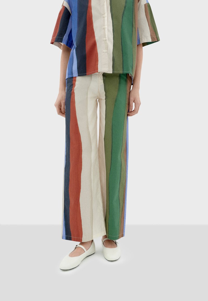Gestreiftes Baumwoll-Outfit, das aus einem kurzärmligen Hemd mit Knöpfen und einer weiten Hose in Blau-, Grün-, Rot- und Beigetönen besteht, kombiniert mit weißen Schuhen.