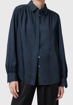 Kobieta w ciemnogranatowej koszuli na guziki z długim rękawem o subtelnym fakturowanym wzorze oraz czarnych spodniach, stojąca na jednolitym tle.