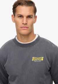 Joven con el cabello corto que lleva una sudadera gris con un logo amarillo de "Superdry Supply Co." en el pecho, mirando hacia adelante.
