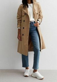 Person som bär en beige trenchcoat, vit sweatshirt, blå jeans och vita sneakers, stående med ena handen i fickan mot en neutral bakgrund.