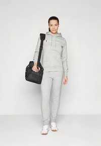 Γυναίκα που φοράει γκρίζα φόρμα Champion και παντελόνια φόρμας, λευκά αθλητικά παπούτσια, κρατώντας μια μαύρη τσάντα γυμναστικής στον ώμο της, στεκόμενη μπροστά σε μια απλή φόντο.