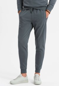 Graue Jogginghosen mit einem Kordelzugbund, Seitentaschen und schmaler Passform, kombiniert mit hellgrauen Sportschuhen. Glatte Textur.