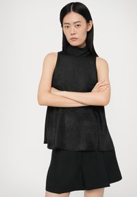 Zwart mouwloos topje met een hoge hals, gemaakt van een gestructureerde stof. Losse pasvorm, gecombineerd met zwarte shorts, zowel minimalistisch als modern.