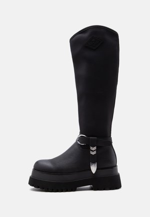 Botte noire haute avec semelle épaisse, bout rond, bride latérale avec boucle argentée et logo REPLAY près du haut sur la partie supérieure lisse.
