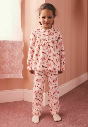 Fetiță tânără stând în interior, purtând pijamale albe cu modele de desene animate roșii, pereți roz, perdea de dantelă la fereastra din stânga ei.