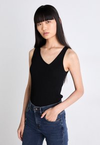 Svart ribbet tanktopp med V-hals og brede skulderstropper, brukt med blå høyt midje jeans, som viser en figursydd silhuett.