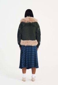 Giacca bomber verde con colletto e orlo in pelliccia sintetica, caratterizzata da dettagli ricamati. Abbinata a una gonna a quadri blu e calzini bianchi.