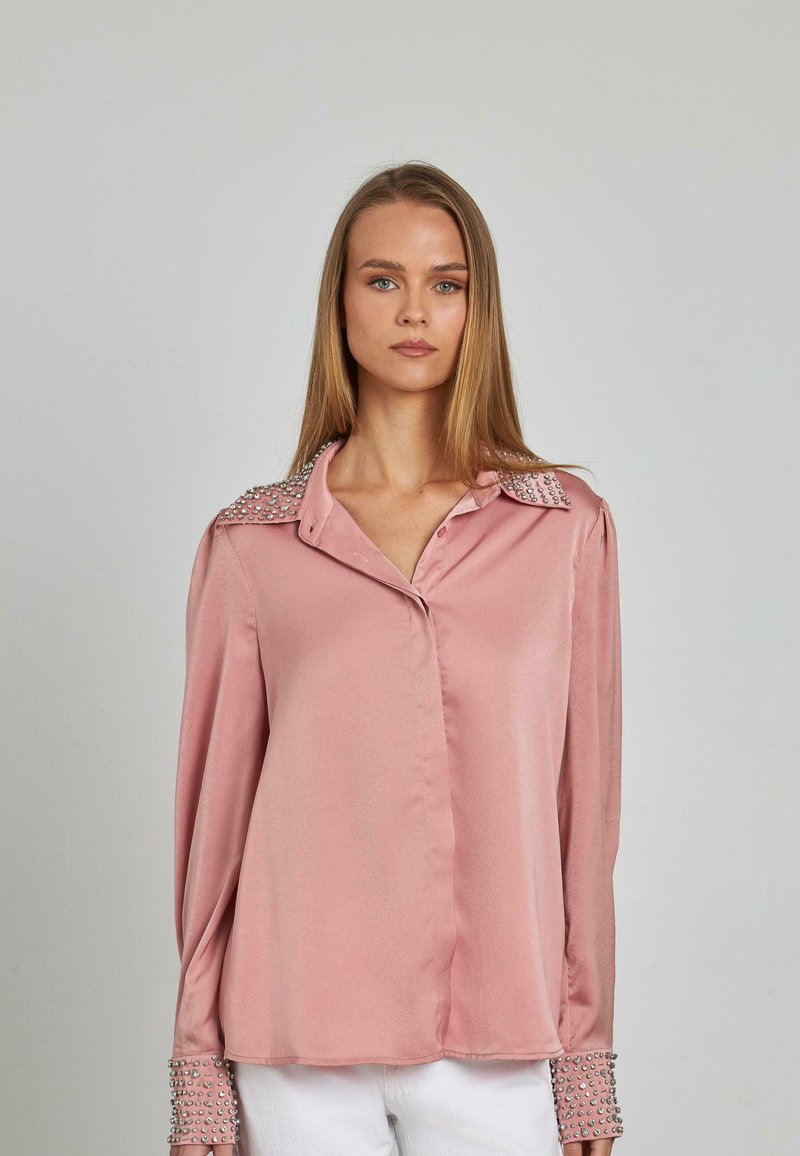 Rosa Satinbluse mit einem Kragen, der mit glänzenden Verzierungen geschmückt ist, sowie verzierten Manschetten. Sie hat einen lockeren Schnitt und lange Ärmel.