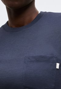 Εγγύς πλάνο ατόμου που φοράει απλό σκούρο μπλε μπλουζάκι με στρογγυλή λαιμόκοψη, τσέπη στο στήθος και μικρή λευκή ετικέτα στη δεξιά πλευρά της τσέπης.