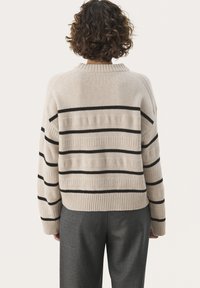 Pull beige rayé avec des lignes horizontales noires, doté d'un col côtelé et d'une maille texturée, associé à un pantalon gris ajusté.