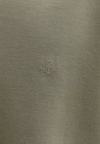 Világos olívazöld kötött anyag kis geometriai logóval, amelyet középen, azonos színű cérnával hímeztek.