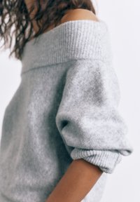 Osoba s kudrnatými tmavými vlasy, která nosí světle šedý svetřík s odhalenými rameny a vyhrnutými rukávy, na jednobarevném bílém pozadí.