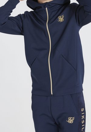 Marineblå hættetrøje med lynlås, tekstureret stof, gylden lynlås og logo. Har sidelommer og hætte, parret med matchende bukser.