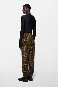 Μαύρο τοπ με μακριά μανίκια και εφαρμοστή σχεδίαση, συνδυασμένο με φαρδιά παντελόνια σε καφέ και χρυσό χρώμα με σχέδια, που διαθέτουν ελαστική μέση και πιεσμένα μανίκια.