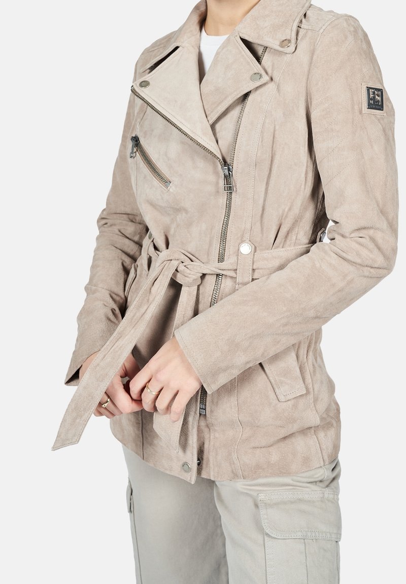 Femme portant une veste de motard beige en daim ceinturée, avec une poche poitrine zippée et des détails à boutons-pression, associée à un pantalon cargo clair.