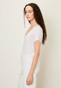 Balta trumpa rankovėmis V formos iškirptė viršutinė dalis, pagaminta iš lengvos medžiagos, pasižyminti laisvu siluetu ir šiek tiek pailgintu hemu, kad sukurtų atsitiktinį įvaizdį.