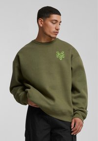 Olijfgroene sweatshirt met een ronde hals en een losse pasvorm. Heeft een felgroen geborduurd logo op de borst. Gemaakt van een katoenmix.