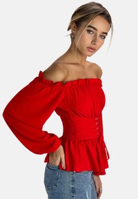 Blusa rossa a spalle scoperte con bordi arricciati, maniche lunghe, vita aderente con arricciature e orlo peplo. Abbinata a jeans blu.