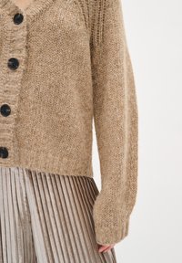 Beżowy sweter z dzianiny z dekoltem w serek, z pięcioma czarnymi guzikami, teksturalną przędzą i ściągaczami, w połączeniu z plisowaną metaliczną spódnicą.