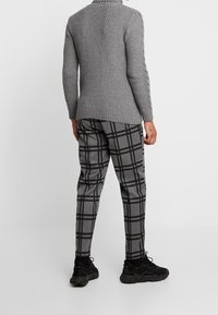 Personne portant un pull gris en tricot texturé et un pantalon à carreaux gris avec des baskets noires, debout devant un fond blanc uni.
