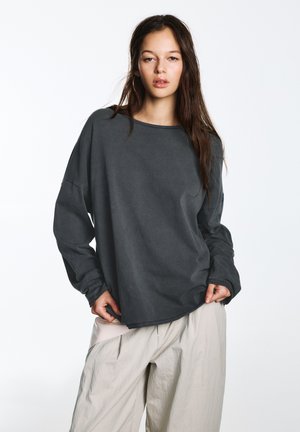 Sivá košeľa s dlhým rukávom s voľným strihom, okrúhlym výstrihom a viditeľným prešívaním. Kombinovaná s ľahkými, textúrovanými, voľne sfarbenými nohavicami.