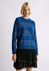 Suéter de punto azul con rayas horizontales, que presenta una textura suave y acentos brillantes, combinado con una falda negra de volantes.