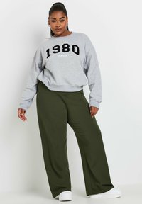 Grå sweatshirts med "1980 PARIS" tryckt i svart, i kombination med olivgröna vida byxor. Modellen bär vita skor med en slät yta.
