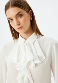 Blusa blanca de manga larga hecha de una tela suave, con un gran cuello con volantes y un cierre de botón en el cuello. Diseño minimalista.