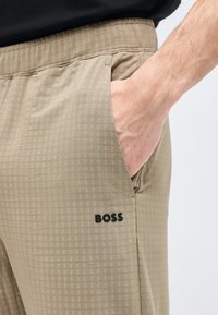Χέρι στην τσέπη μπεζ καρό παντελονιού με ελαστική μέση και μαύρο λογότυπο "BOSS" στο πλάι.