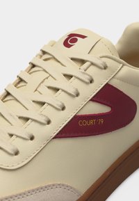 Záber zblízka na krémovú tenisku s béžovými šnúrkami, bordovým bočným detailom, nápisom "COURT '79" a hnedou podrážkou.