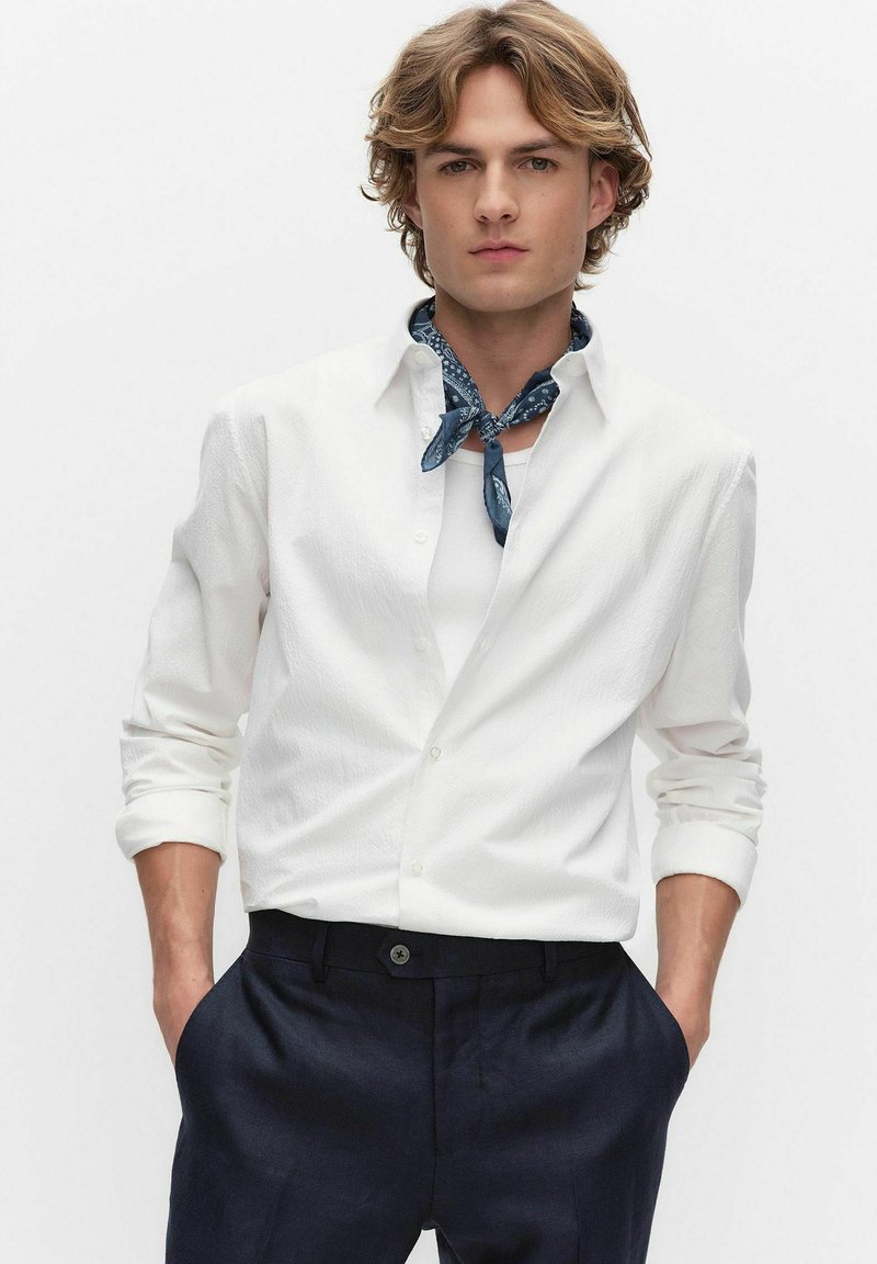 Jonge man met een wit overhemd over een wit hemd zonder mouwen, marineblauwe broek en een blauw halsdoek met patroon, met handen in de zakken.