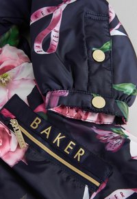 Blumenbedruckte Steppjacke aus Satin mit navy Hintergrund, goldenen Akzenten und einem auffälligen goldenen Reißverschluss mit "BAKER"-Logo.