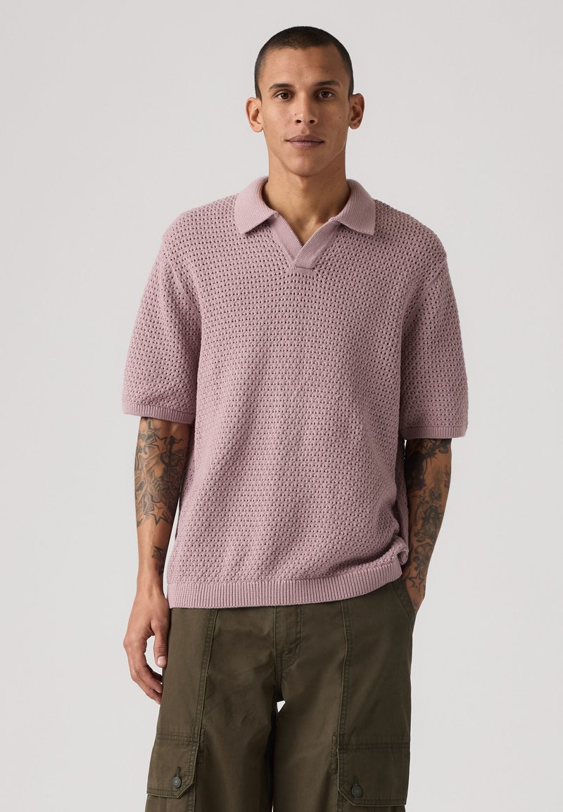 Homme aux cheveux courts portant un polo en maille violet clair à manches courtes et un pantalon cargo vert olive, debout les mains dans les poches.