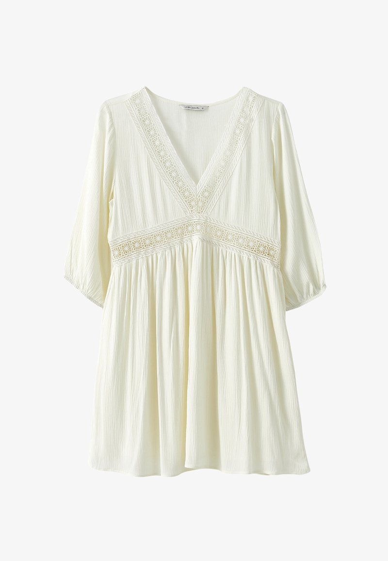 Tunica bianca, leggera e texturizzata con scollo a V, maniche corte e dettagli in pizzo lungo il colletto e in vita. Silhouette fluida.