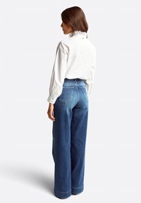 Weiße Bluse mit einer Spitzeneinfassung und langen Ärmeln, kombiniert mit weit geschnittenen blauen Denim-Jeans, die zwei Gesäßtaschen und umgeschlagene Säume haben.