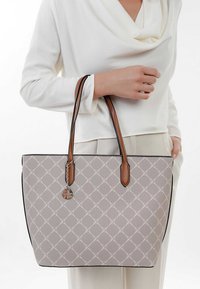 Sac fourre-tout en tissu gris clair avec un motif en losange, poignées en cuir marron et un charme argenté. Présente des bordures noires et un design structuré.