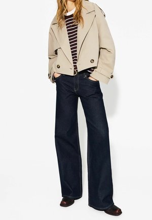 Kvinde iført beige cropped jakke, sort- og hvidstribet skjorte, mørke vidde jeans og mørke sko med hænderne i lommerne.