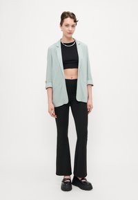 Hellgrüner Blazer über einem schwarzen Crop-Top, kombiniert mit schwarzen Schlaghosen und klobigen schwarzen Plateauschuhen, mit glatter Textur und minimalistischem Design.