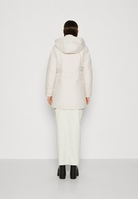 Parka blanche matelassée avec capuche, présentant une coupe droite et des manches longues, associée à une longue jupe blanche et des bottes à plateforme noires.