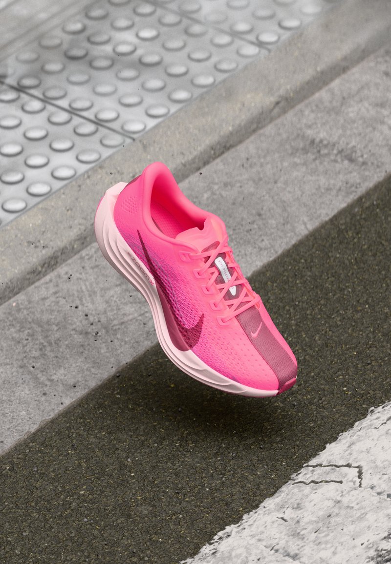 Nike urheilujalkineet kirkkaan vaaleanpunaisina, joissa on verkkopäällinen, pehmustettu kaulus, valkoinen välikerroksen ja kontrastoiva tummempi vaaleanpunainen varvaskuppi sekä Swoosh-yksityiskohta.